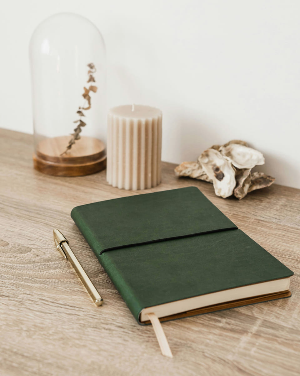 A green linen-bound planner and a brass pen on an oak desk, beside a ribbed candle.