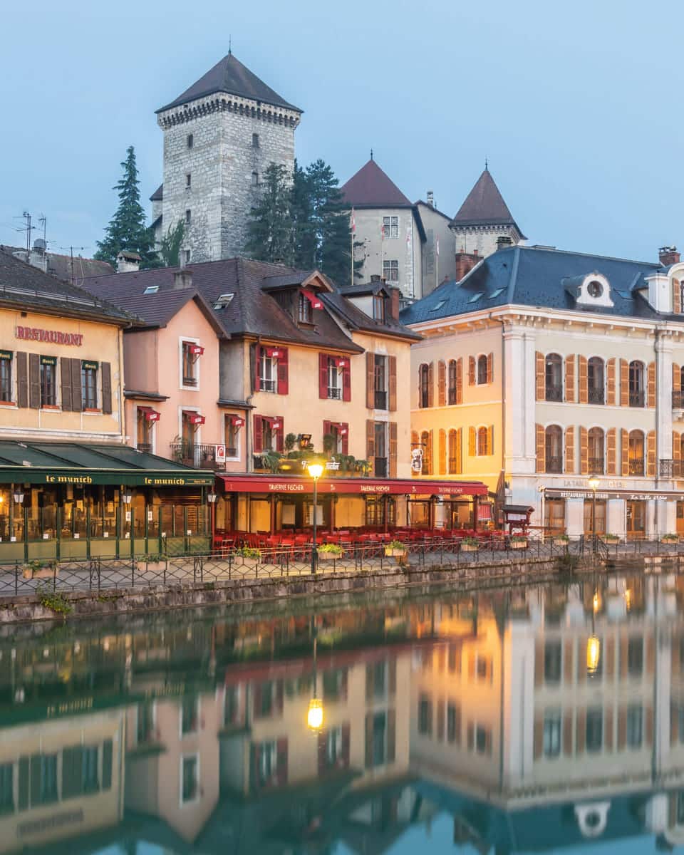 The Château d’Annecy rising above the pastel façades and red shutters of the quai Perrière, mirrored in still canal water.