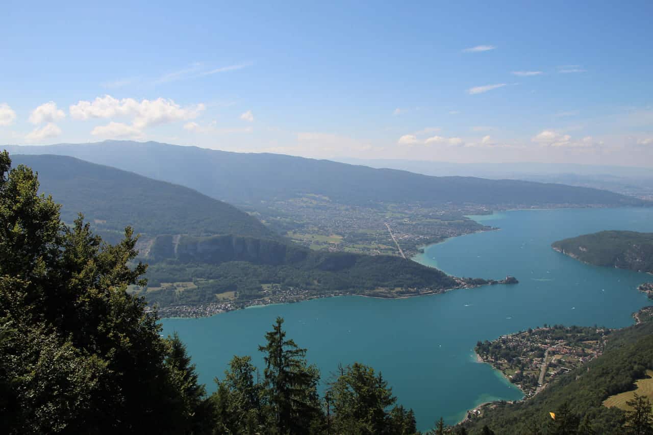 Lake Annecy seen from the wooded slopes above, turquoise water between green mountains under a high summer sky.