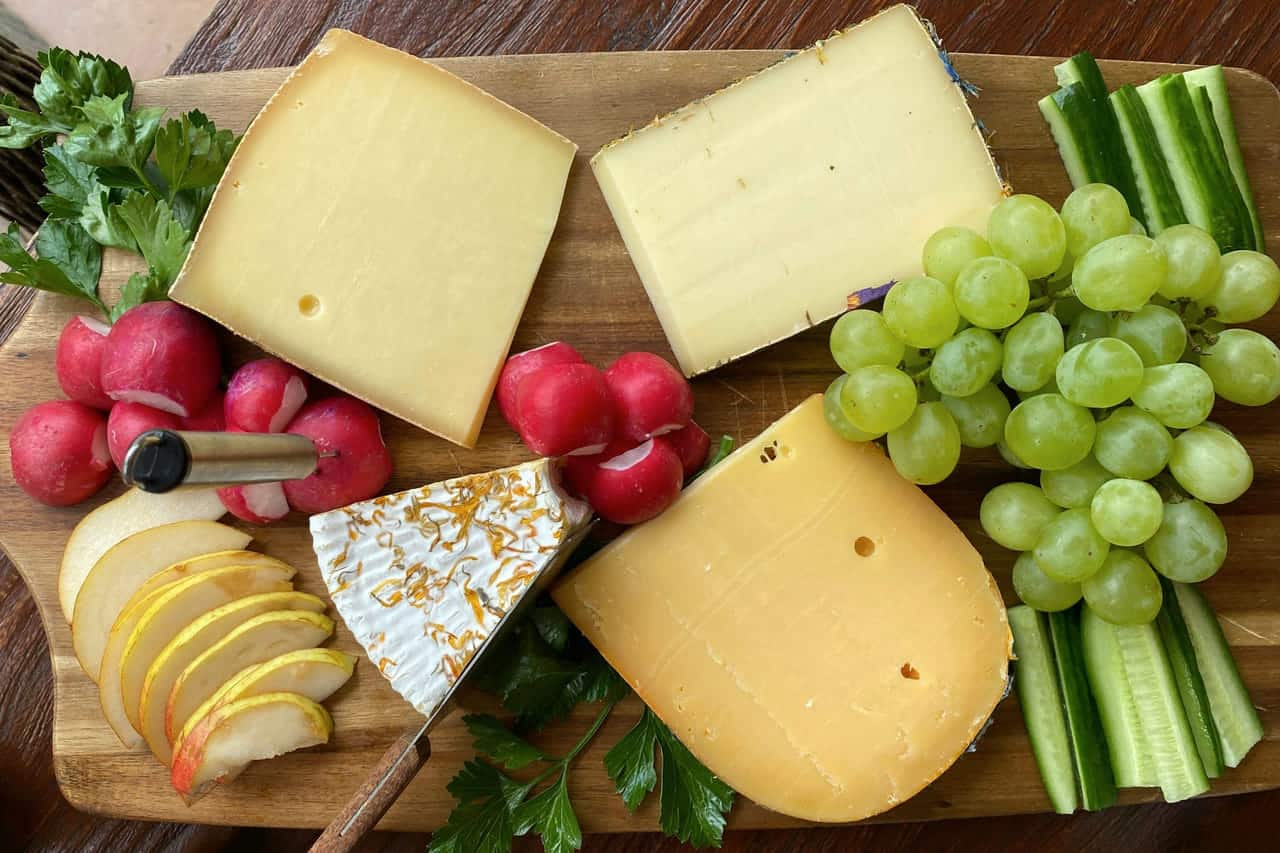 Wedges and blocks of firm yellow cheese on a wooden board with green grapes, celery sticks and sliced radishes.