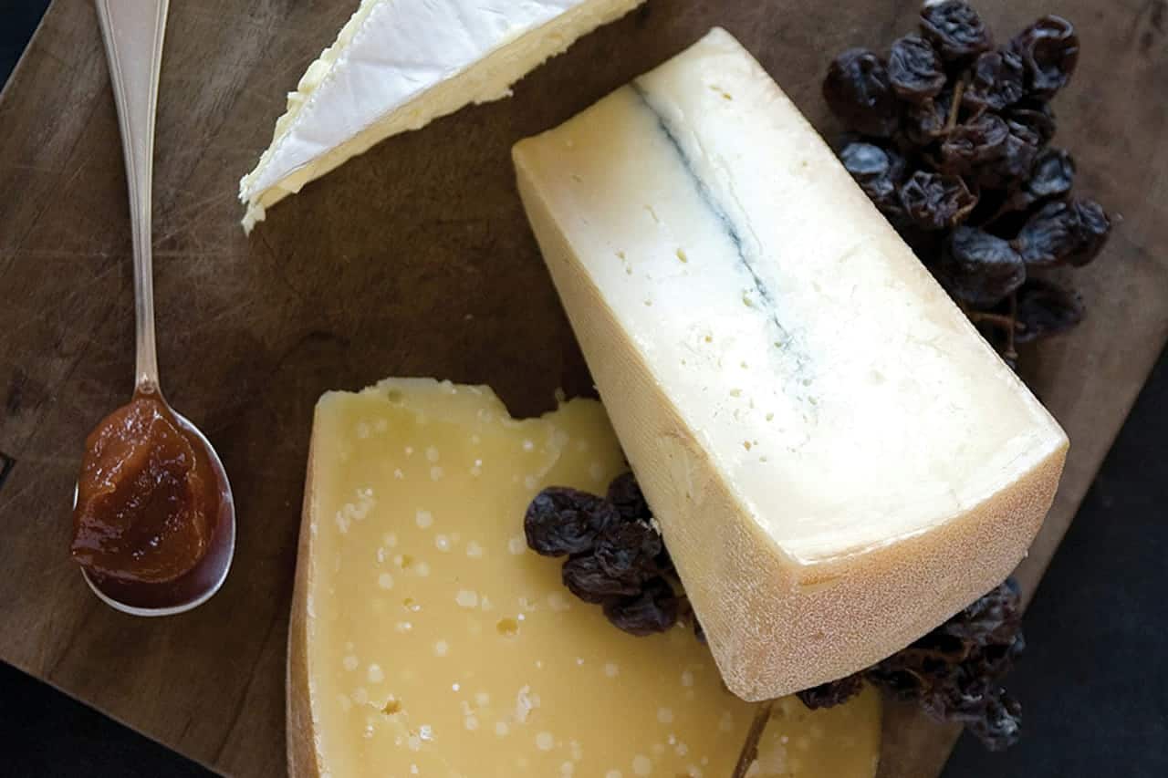 A wedge of white-rind soft cheese, a wedge of semi-soft cheese with a fine dark line through its centre and a piece of firm mountain cheese on a worn wooden board, with dried grapes and a spoonful of fruit paste, on a dark ground.