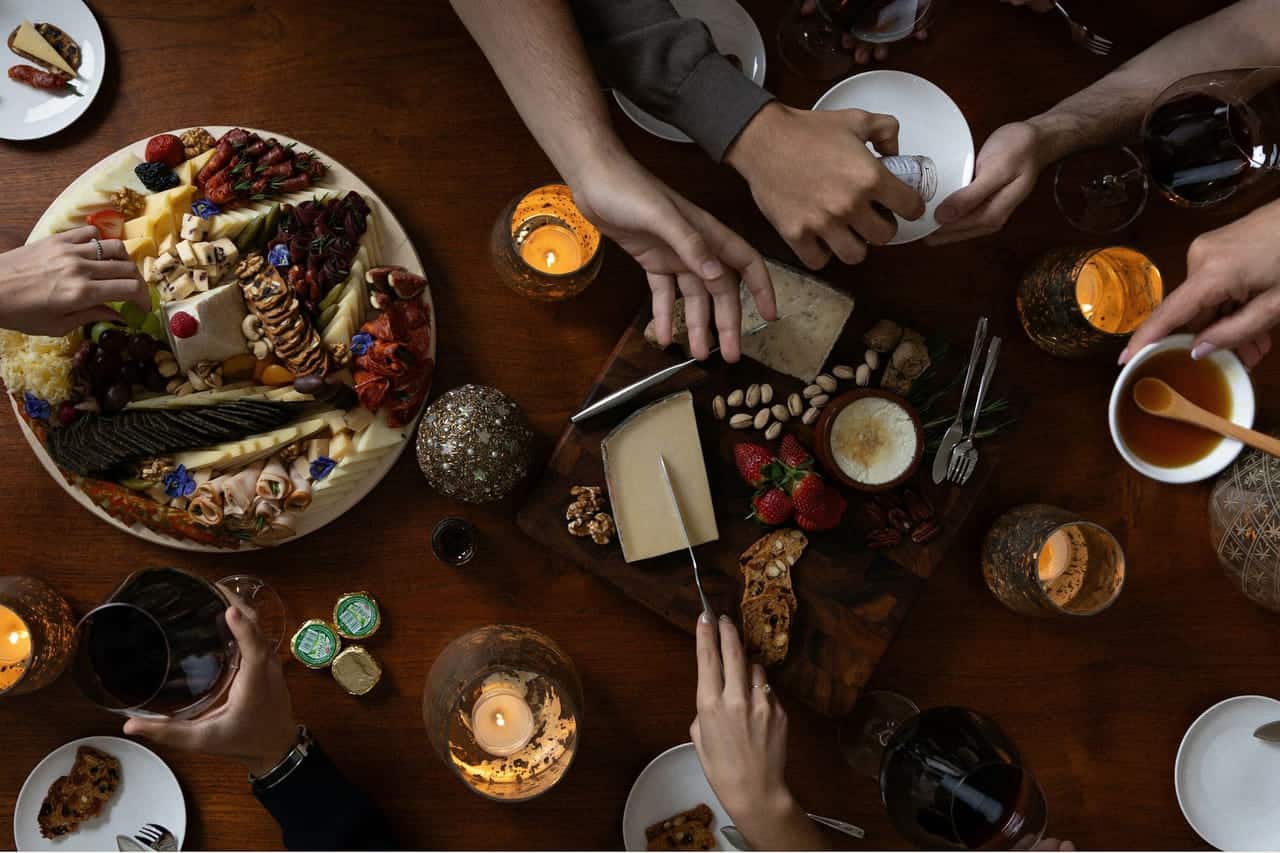 Several pairs of hands reaching across a dark wooden table set with a charcuterie board, small plates, cups and candles.