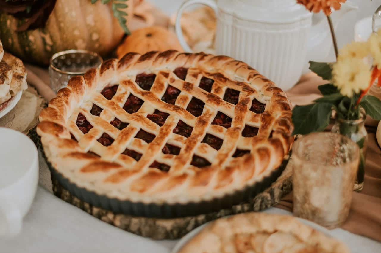 A lattice-top berry pie on a wooden round beside an apple galette, a white teapot, small pumpkins and dahlias on a linen-covered table.