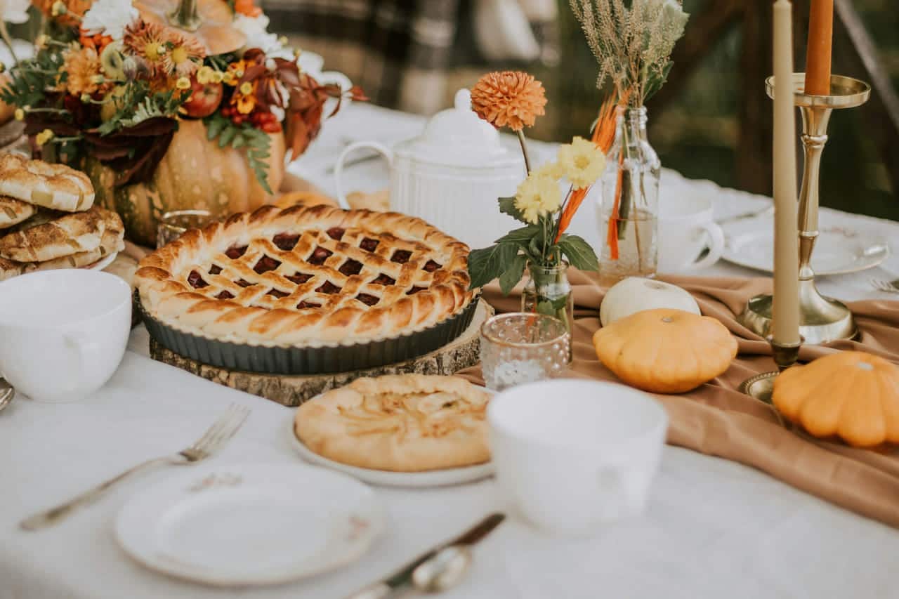 An autumn table outdoors dressed in white linen and a caramel runner: a lattice-top pie, an apple galette, a teapot, small pumpkins, orange and ivory tapers in brass holders and a centerpiece of dahlias in a pumpkin, with a plaid blanket over a chair.