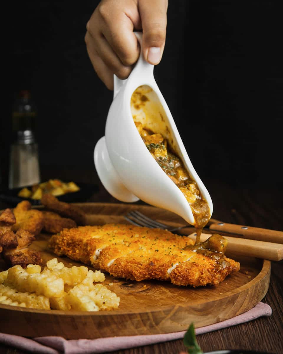 A hand pouring mushroom gravy from a white gravy boat over a crisp breaded chicken cutlet on a wooden board, against a dark background.