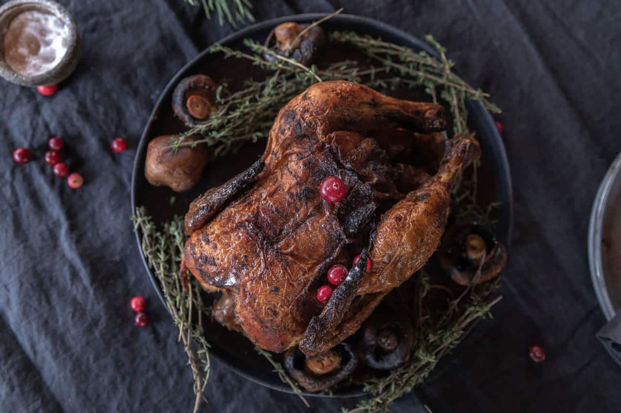 Overhead view of a whole roast bird scattered with cranberries, on a dark plate ringed with thyme and roasted mushrooms, on a charcoal linen cloth with pine sprigs and a stoneware plate.