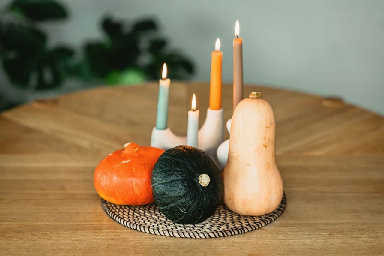 Taper candles of different heights in pale holders beside a small orange pumpkin, a dark green squash and a butternut squash on a woven mat on a round wooden table.