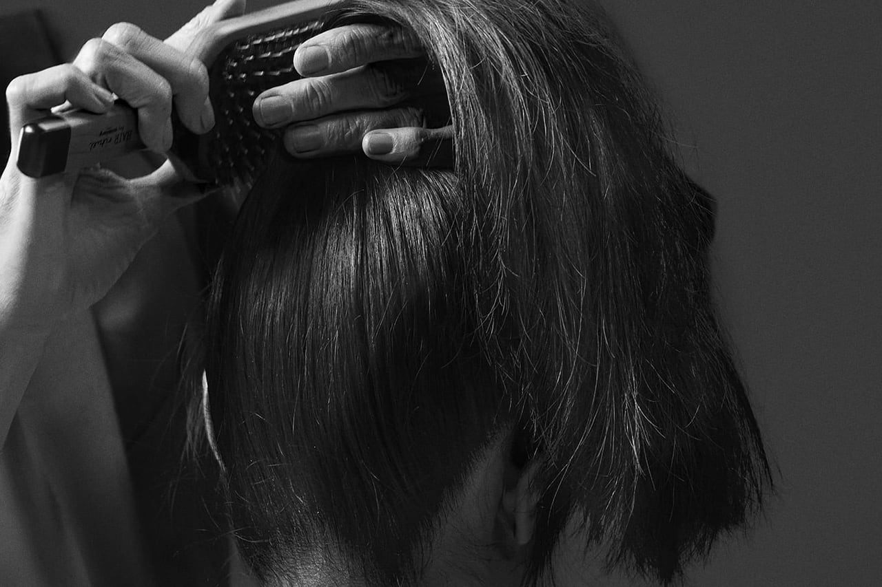 Seen from behind, a hand runs a Sisley paddle brush back through a dark bob, in black and white.