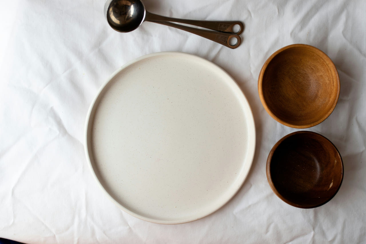 Overhead view of a matte cream stoneware plate beside two wooden bowls and brass measuring spoons on white linen.