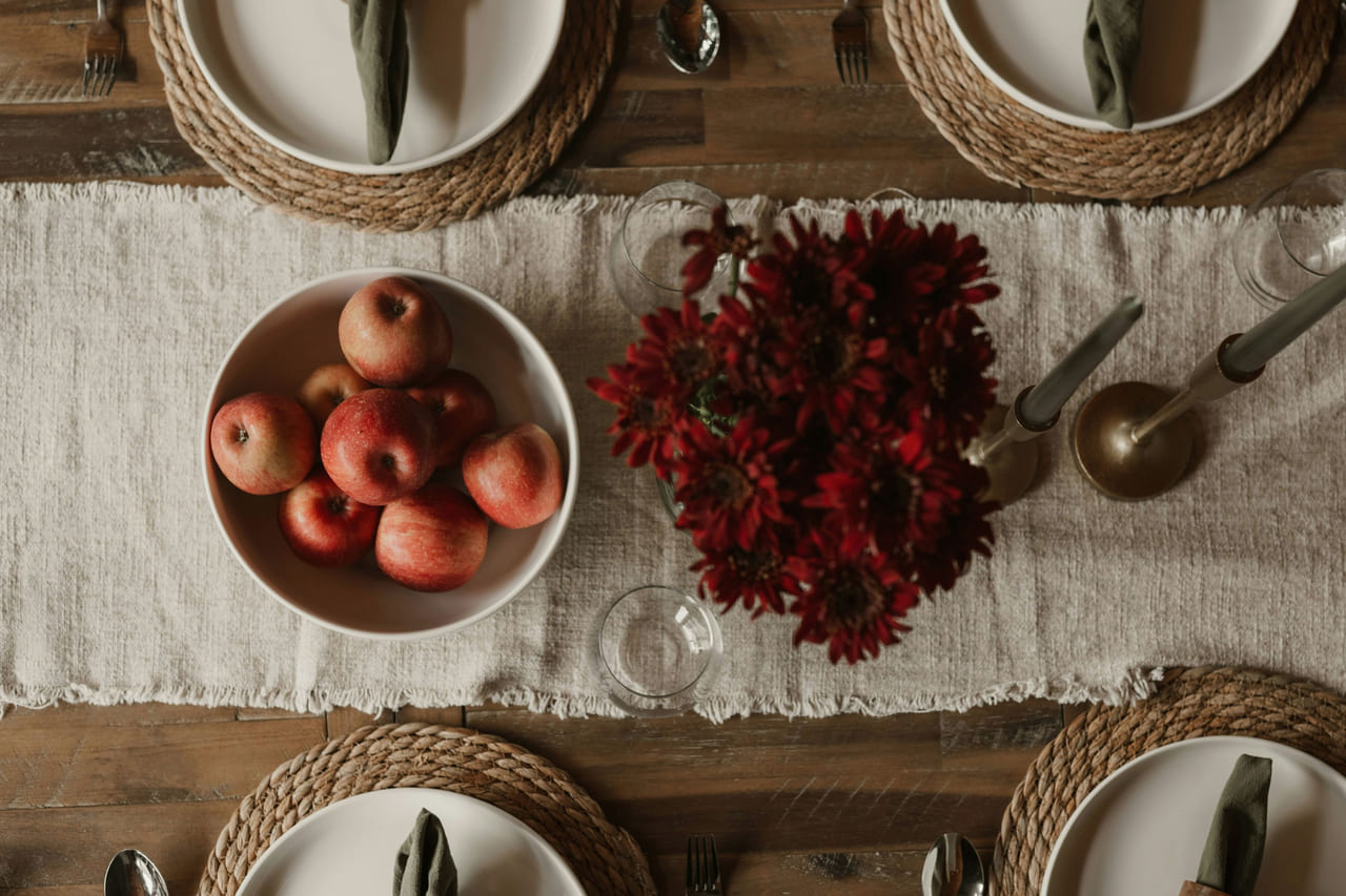 Overhead view of a long table dressed with a linen runner, woven chargers, a bowl of red apples and dark red flowers.